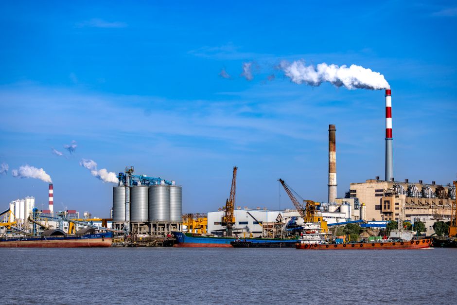 Sustentabilidade — An industrial port scene with smokestacks, silos, and cranes by the water, showcasing urban industry.